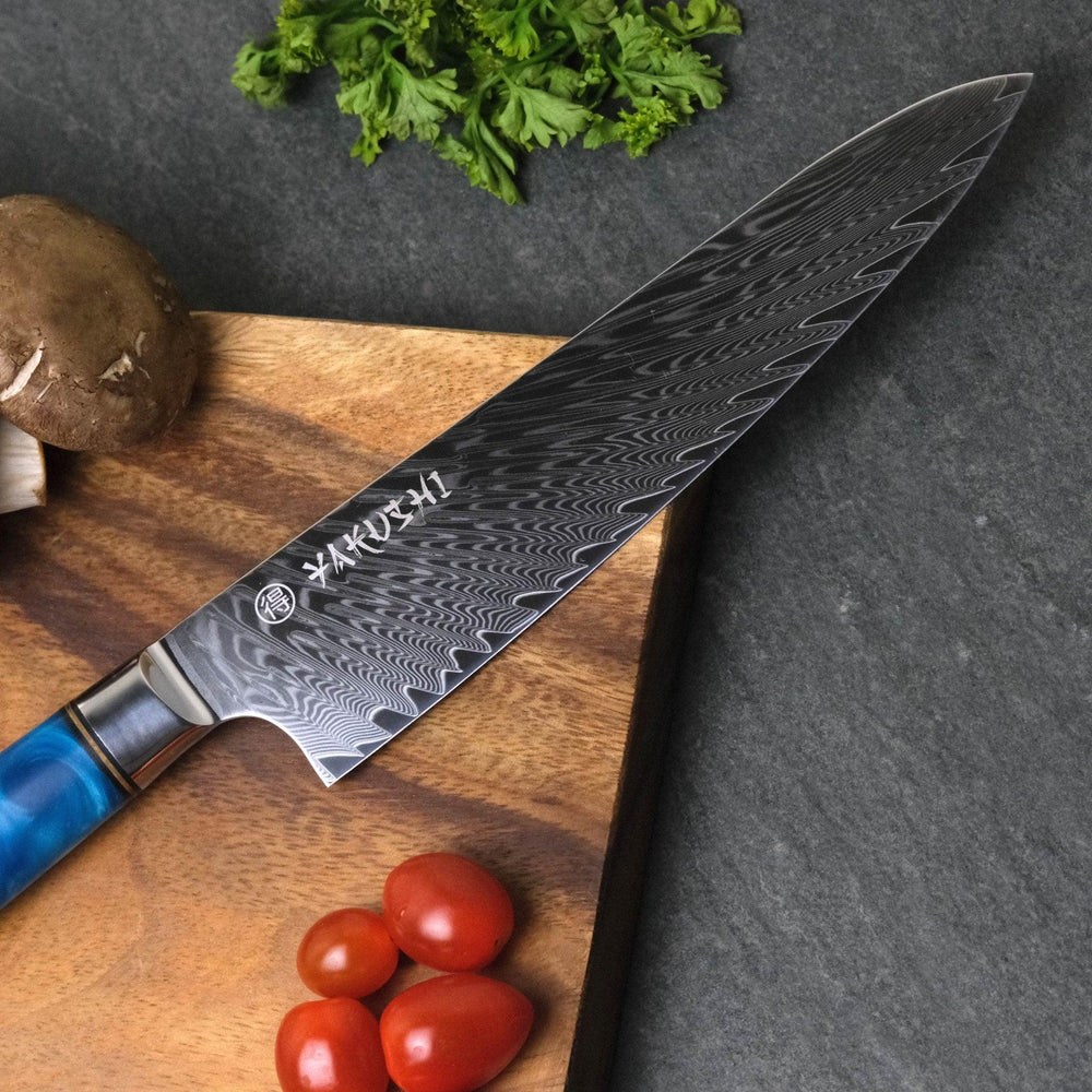 Close view of the layered pattern on a Yakushi Damascus chef knife beside tomatoes and parsley